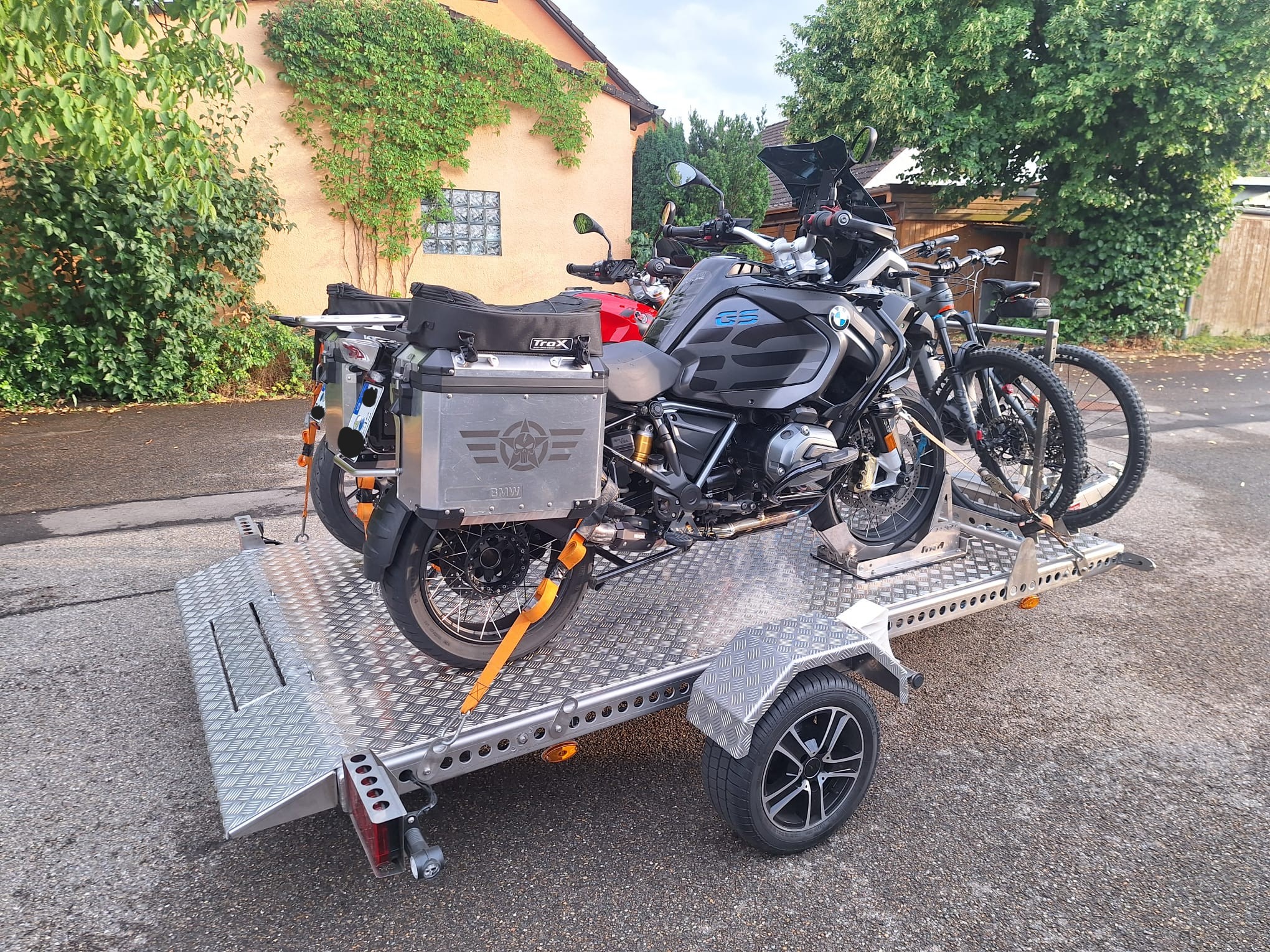 Aluminium Absenkanhänger mit schwarzen Alufelgen und zwei Motorrädern beladen hinter einem VW-Passat auf einer Straße vor einem Haus