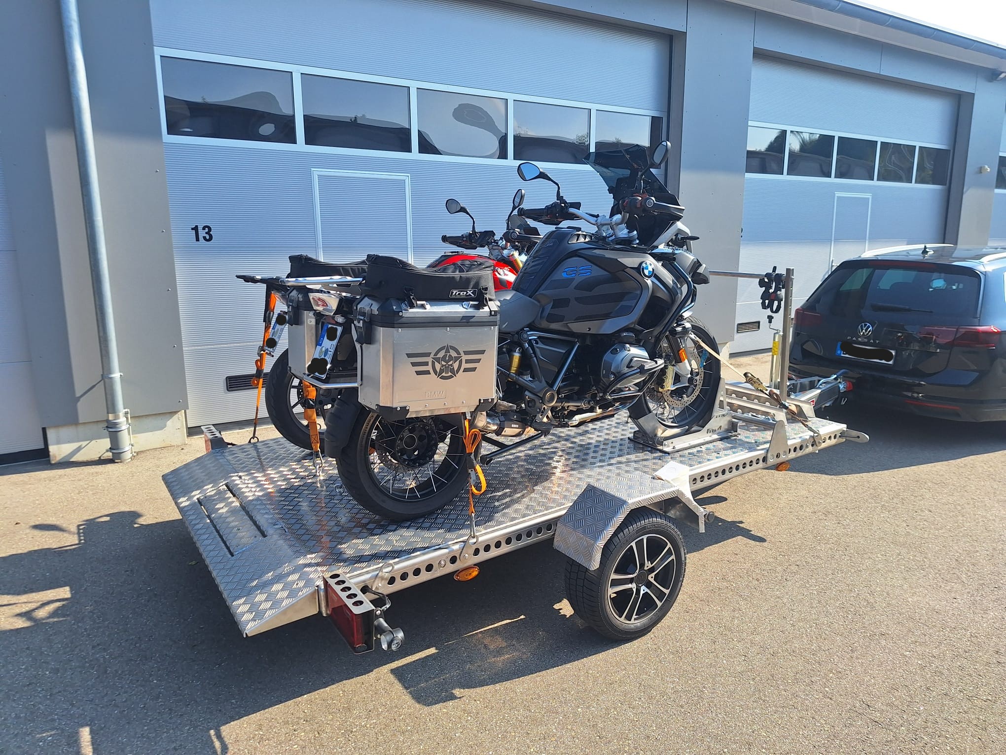 Aluminium Absenkanhänger mit schwarzen Alufelgen und zwei Motorrädern beladen hinter einem VW-Passat vor einem Hallentor