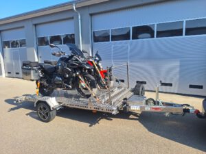 Aluminium Absenkanhänger mit schwarzen Alufelgen und zwei Motorrädern beladen hinter einem VW-Passat vor einem Hallentor