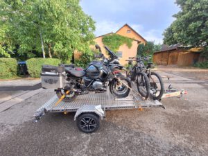 Aluminium Absenkanhänger mit schwarzen Alufelgen und zwei Motorrädern beladen hinter einem VW-Passat auf einer Straße vor einem Haus