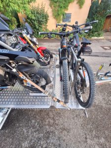 Aluminium Absenkanhänger mit schwarzen Alufelgen und zwei Motorrädern beladen hinter einem VW-Passat auf einer Straße vor einem Haus