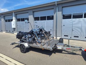 Aluminium Absenkanhänger mit schwarzen Alufelgen und zwei Motorrädern beladen hinter einem VW-Passat vor einem Hallentor