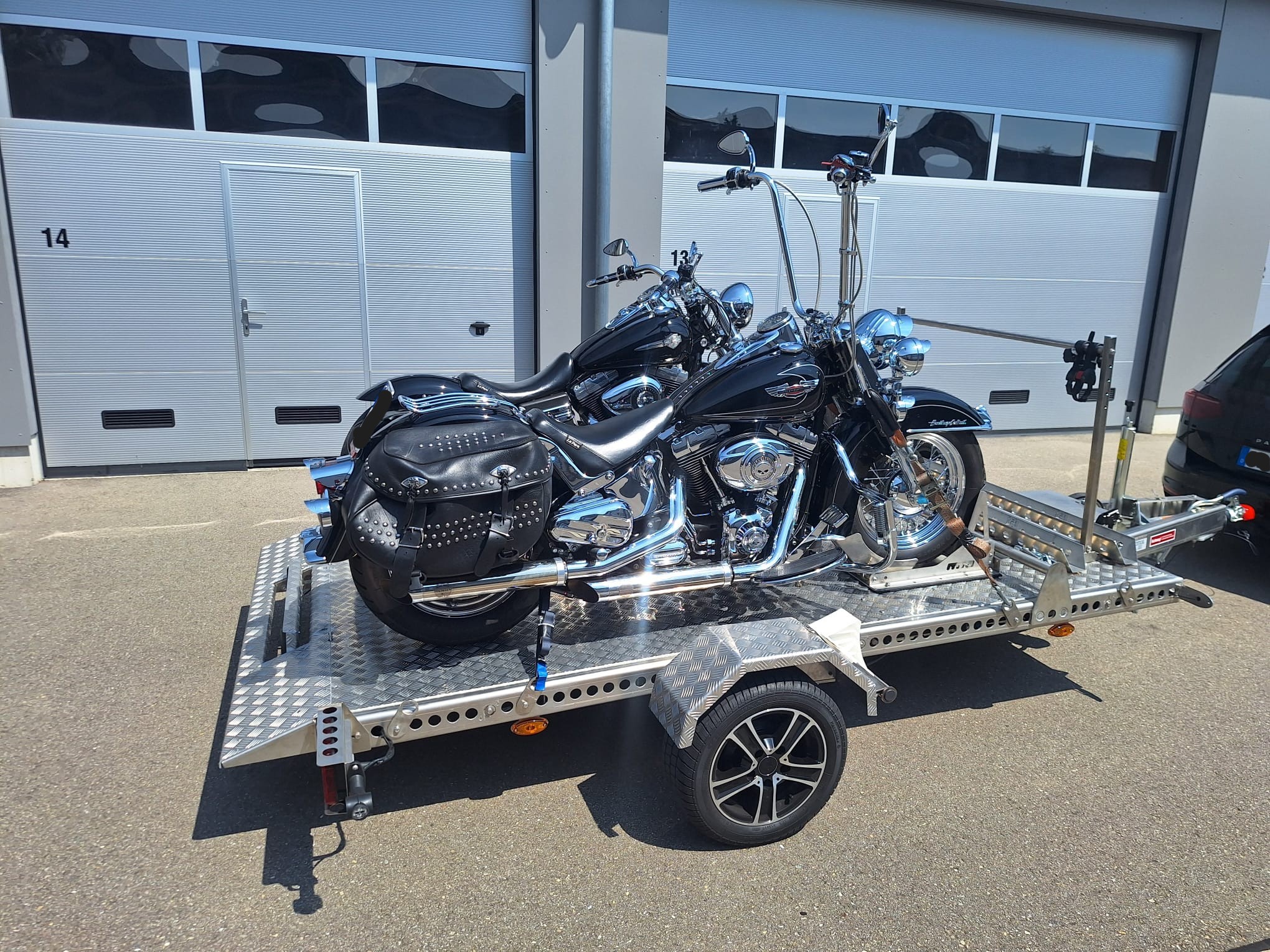 Aluminium Absenkanhänger mit schwarzen Alufelgen und zwei Motorrädern beladen hinter einem VW-Passat vor einem Hallentor