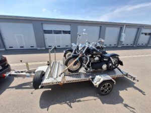 Aluminium Absenkanhänger mit schwarzen Alufelgen und zwei Motorrädern beladen hinter einem VW-Passat vor einem Hallentor