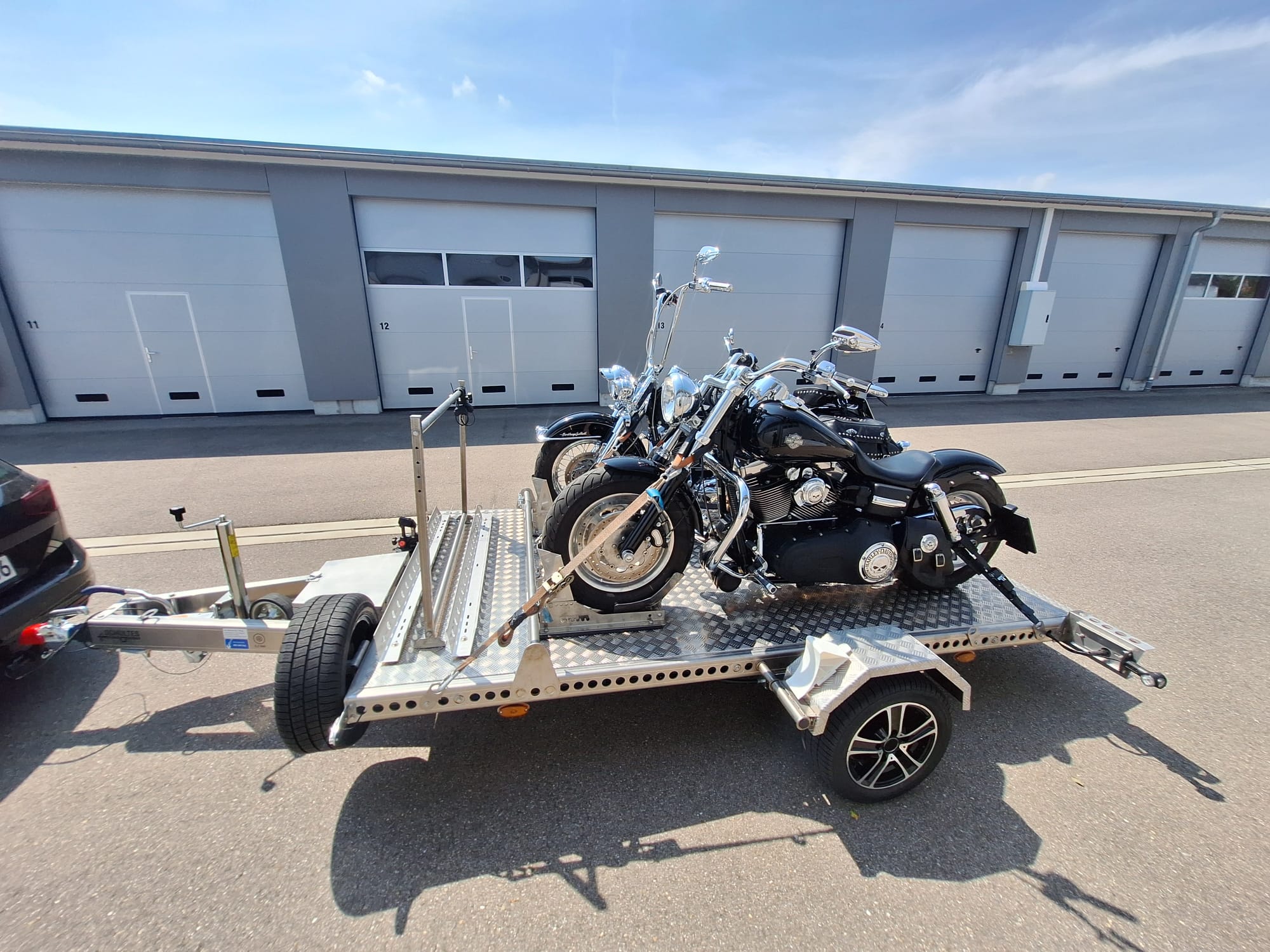 Aluminium Absenkanhänger mit schwarzen Alufelgen und zwei Motorrädern beladen hinter einem VW-Passat vor einem Hallentor