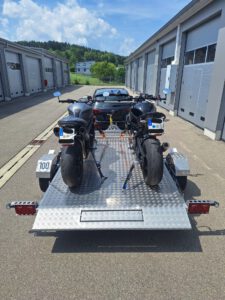 Aluminium Absenkanhänger mit schwarzen Alufelgen und zwei Motorrädern beladen hinter einem VW-Passat vor einem Hallentor