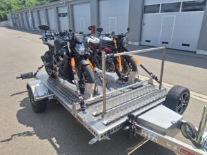 Aluminium Absenkanhänger mit schwarzen Alufelgen und zwei Motorrädern beladen hinter einem VW-Passat vor einem Hallentor