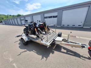 Aluminium Absenkanhänger mit schwarzen Alufelgen und zwei Motorrädern beladen hinter einem VW-Passat vor einem Hallentor