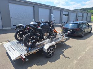 Aluminium Absenkanhänger mit schwarzen Alufelgen und zwei Motorrädern beladen hinter einem VW-Passat vor einem Hallentor