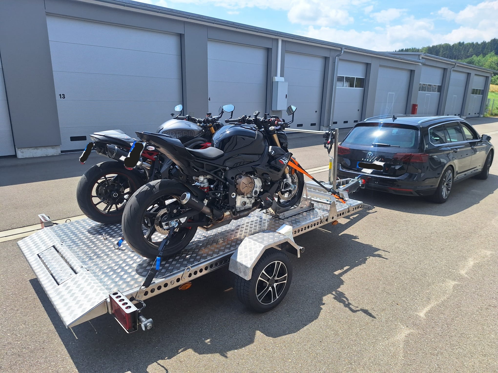 Aluminium Absenkanhänger mit schwarzen Alufelgen und zwei Motorrädern beladen hinter einem VW-Passat vor einem Hallentor