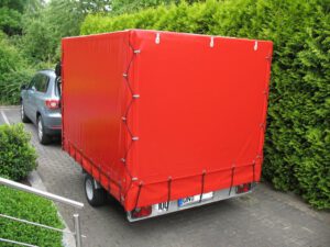 Aluanhänger mit Planengestell und roter Plane vor einer grünen Hecke auf einer Straße