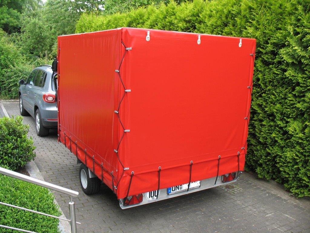 Aluanhänger mit Planengestell und roter Plane vor einer grünen Hecke auf einer Straße