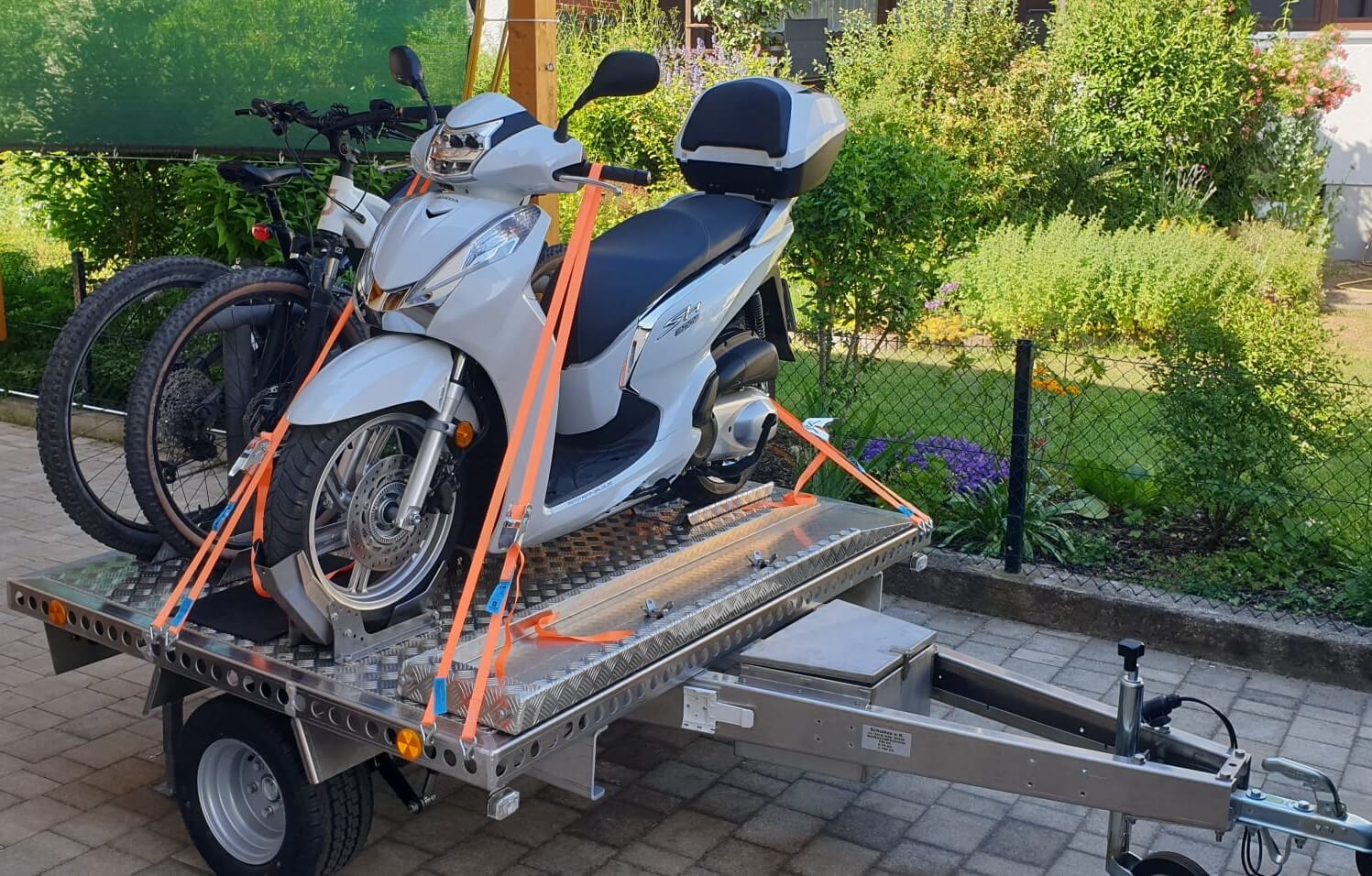 Zwei Fahrräder und ein Roller auf einem Aluminium Plattformanhänger - in einem Hof - von der Seite