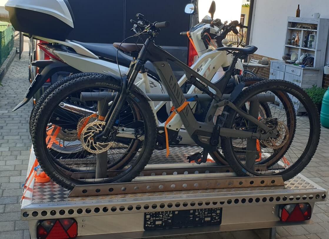 Zwei Fahrräder und ein Roller auf einem Aluminium Plattformanhänger - hinter einem Wohnmobil - von hinten