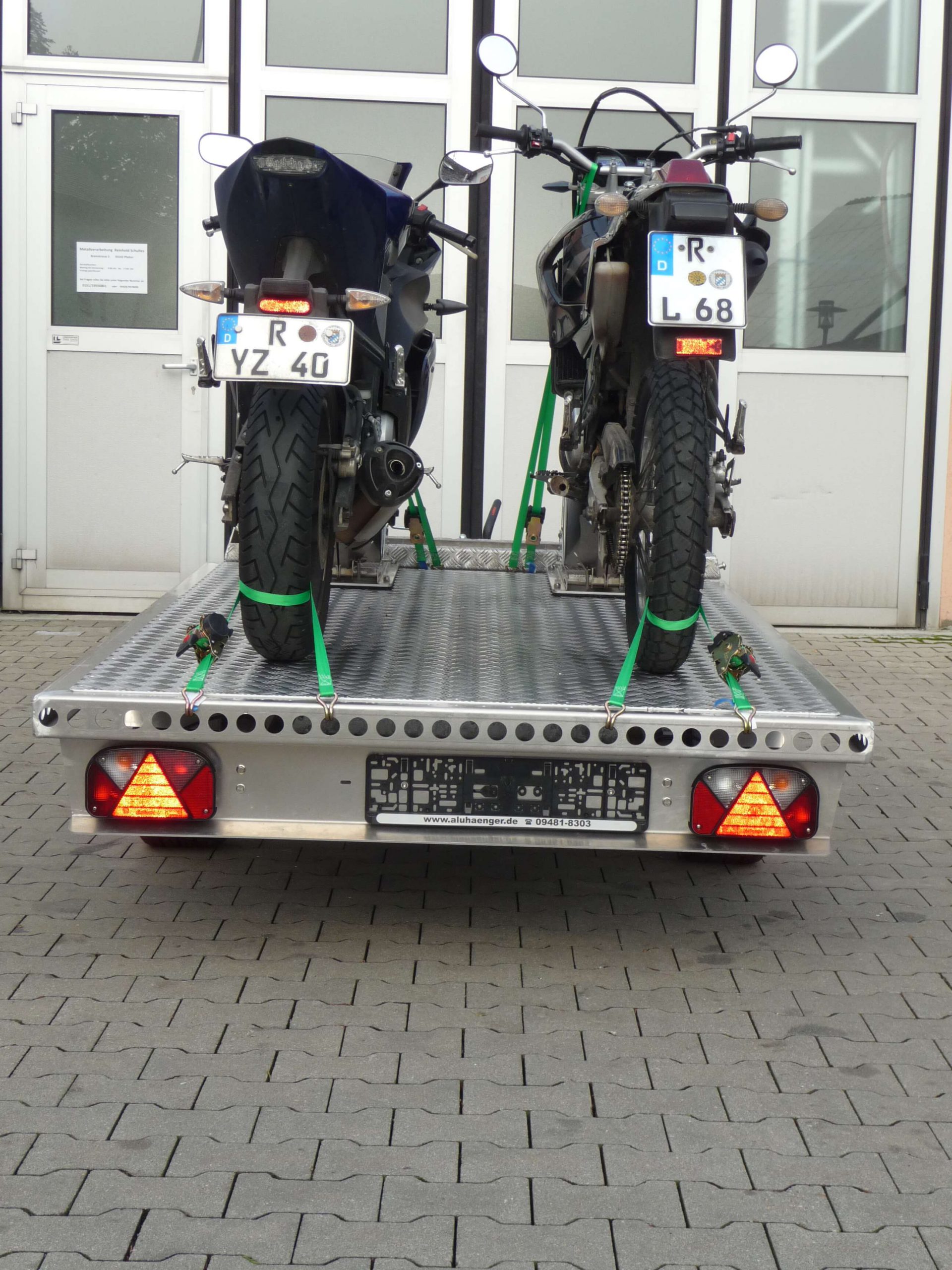 Zwei Motorräder auf einem Aluminium Plattformanhänger vor einem Hallentor - von hinten