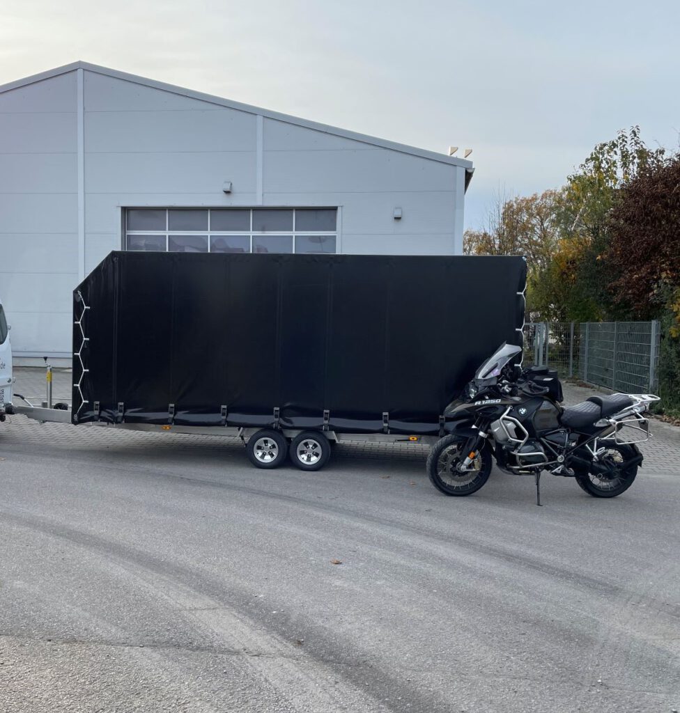 Anhänger mit Hochplane in schwarz mit Motorrad vor einer Halle