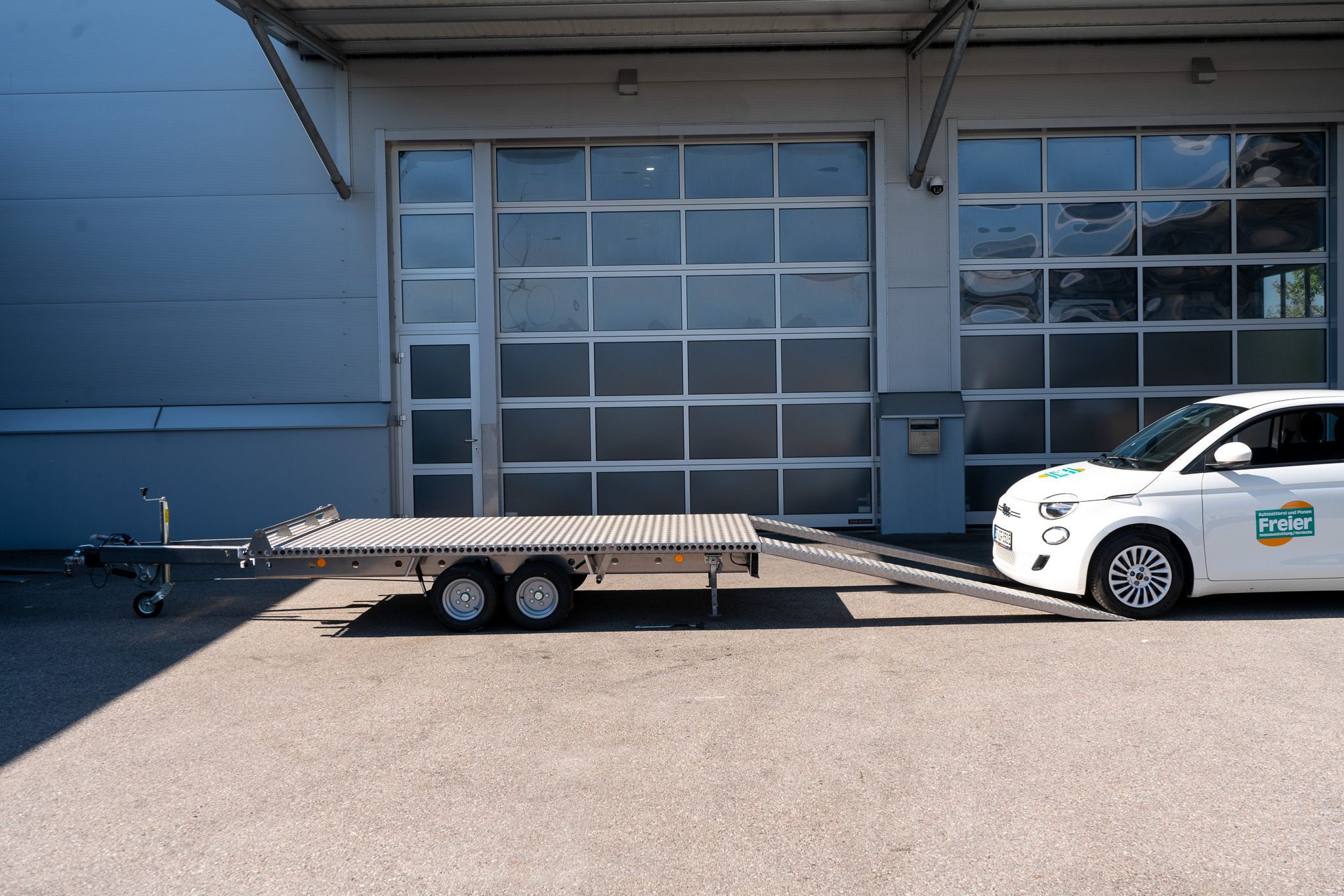 Aluminium Plattformanhänger Tandem, mit einem weißen Fiat 500 hinter dem Anhänger - vor einem Hallentor - Seitenansicht
