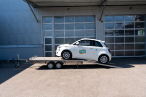 Aluminium Plattformanhänger Tandem, beladen mit einem weißen Fiat 500 vor einem Hallentor - Seite