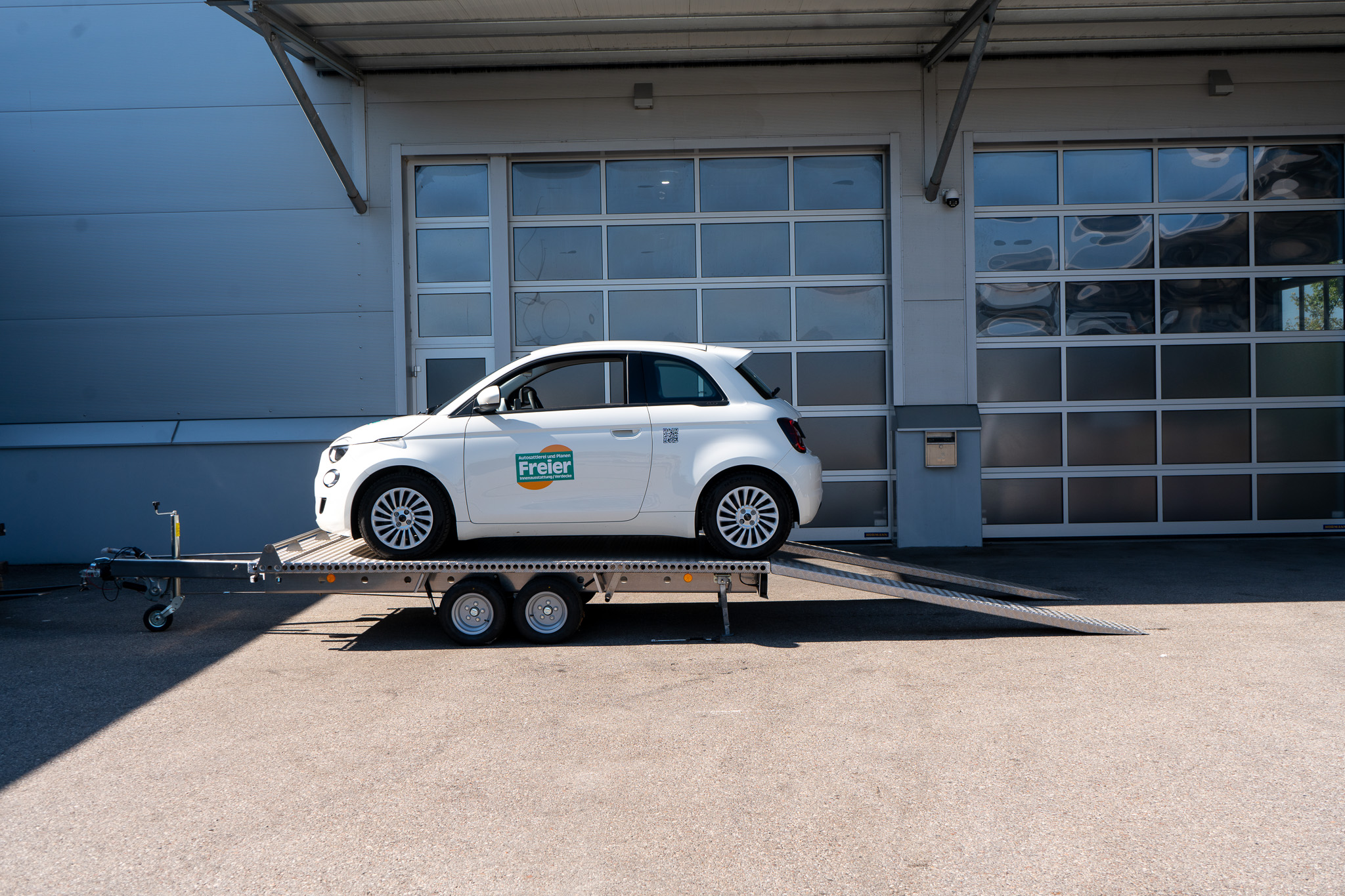 Aluminium Plattformanhänger Tandem, beladen mit einem weißen Fiat 500 vor einem Hallentor - Seite