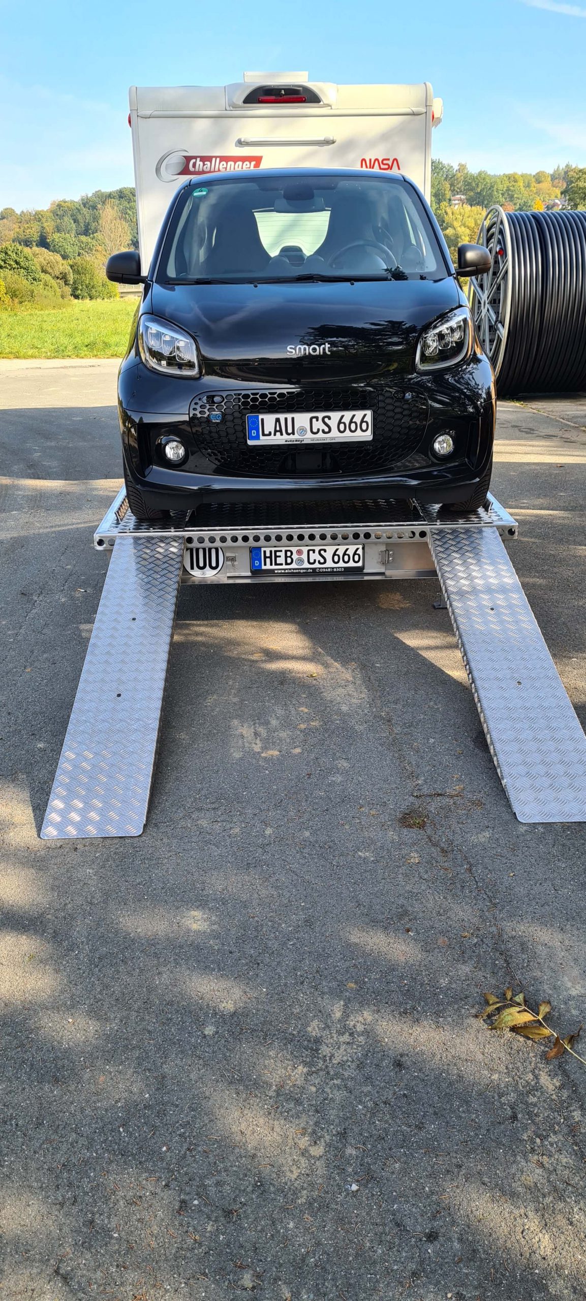 Smart fortwo auf einem Aluminium Plattformanhänger mit Auffahrrampen angehängt an ein Wohnmobil auf einem Parkplatz -Ansicht von hinten