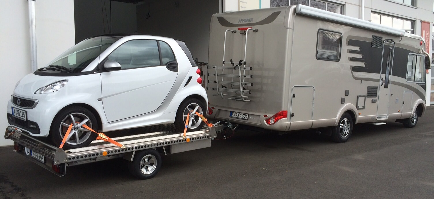 Smart fortwo auf einem Aluminium Plattformanhänger mit Auffahrrampen angehängt an ein Wohnmobil auf einem Parkplatz