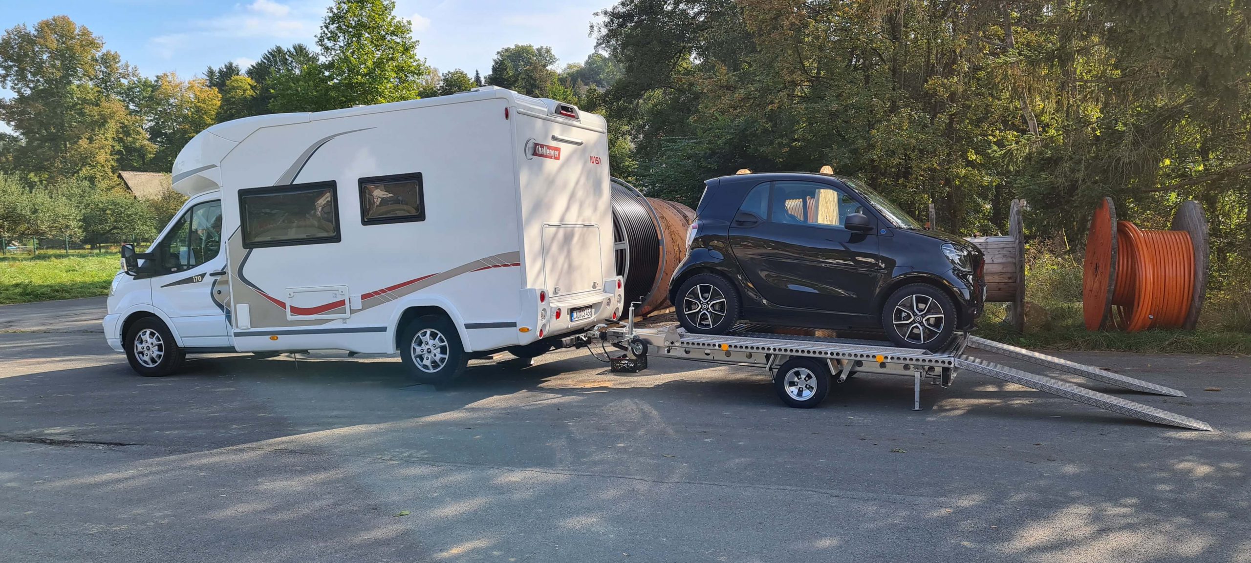 Smart fortwo auf einem Aluminium Plattformanhänger mit Auffahrrampen angehängt an ein Wohnmobil auf einem Parkplatz