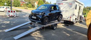 Smart fortwo auf einem Aluminium Plattformanhänger mit Auffahrrampen angehängt an ein Wohnmobil auf einem Parkplatz