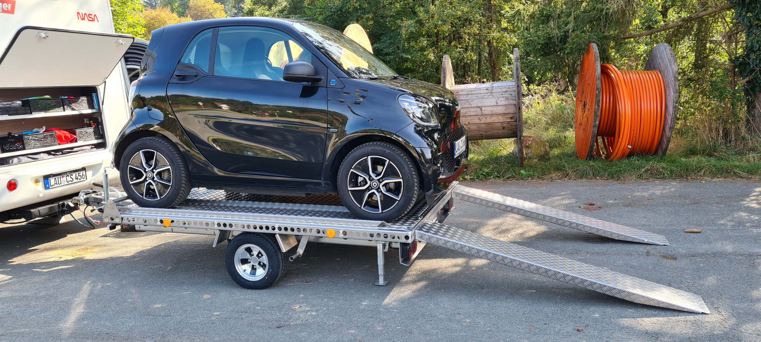 Smart fortwo auf einem Aluminium Plattformanhänger mit Auffahrrampen angehängt an ein Wohnmobil auf einem Parkplatz