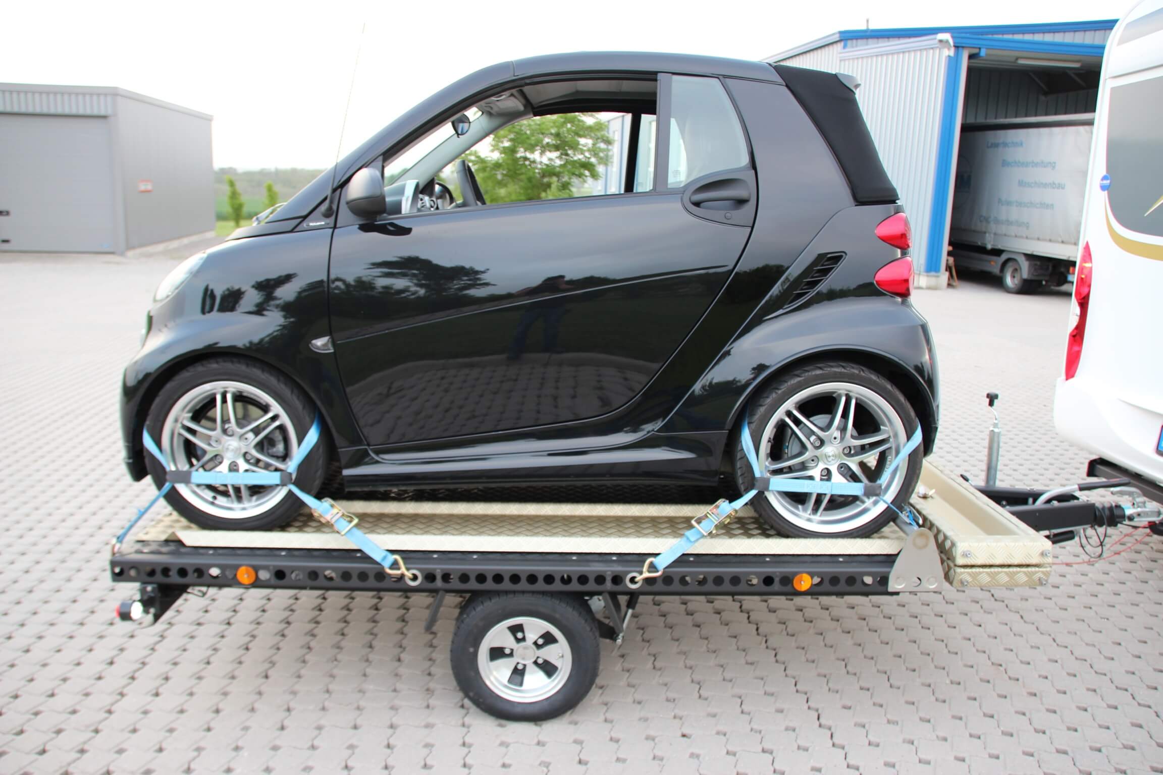 Smart fortwo auf einem Aluminium Plattformanhänger mit Auffahrrampen angehängt an ein Wohnmobil auf einem Parkplatz