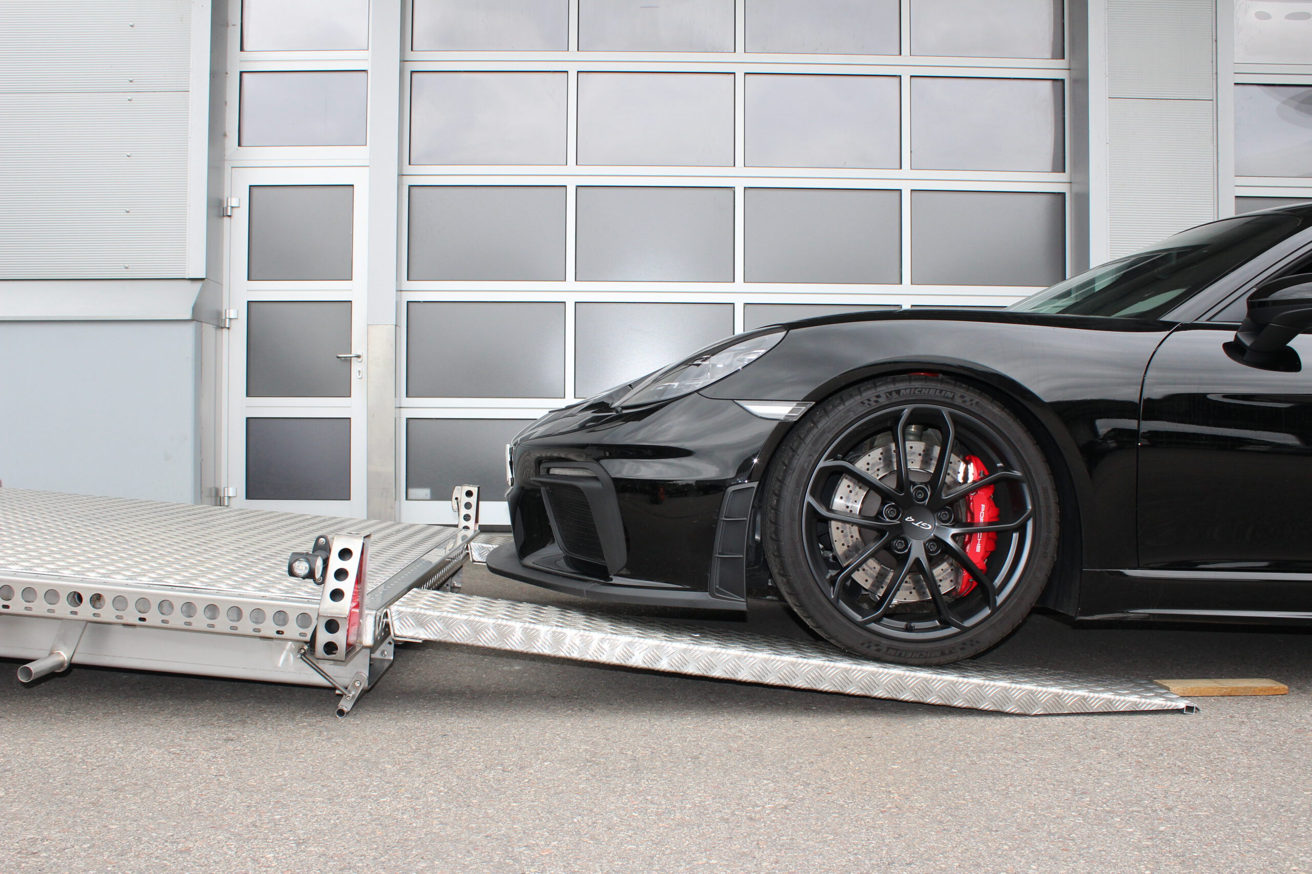 Porsche GT4 fährt über Auffahrrampen auf Tieflader Anhänger - Seitenansicht vor Hallentor