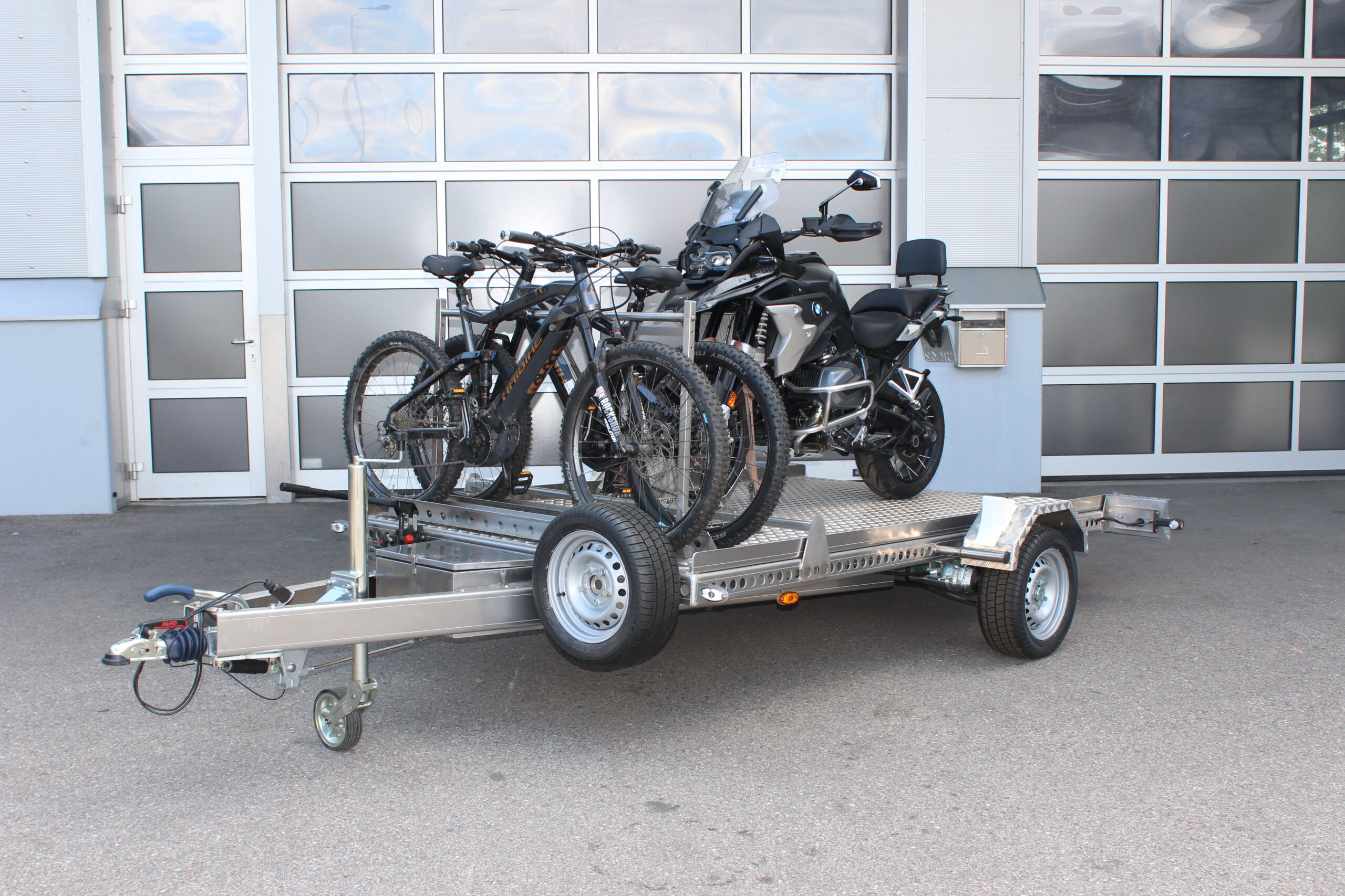 AS300 Aluminium Absenkanhänger in silber mit 2 E-Bikes auf der Plattform vorne quer beladen und mit einer BMW GS beladen vor einem Hallentor