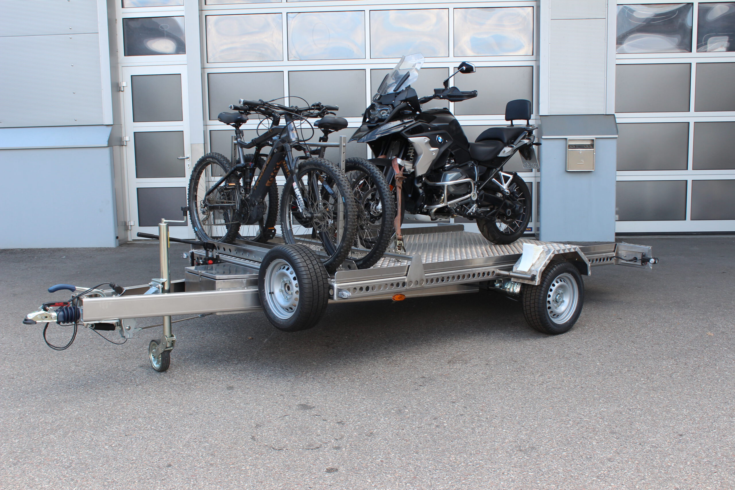AS300 Aluminium Absenkanhänger in silber mit 2 E-Bikes auf der Plattform vorne quer beladen und mit einer BMW GS beladen vor einem Hallentor