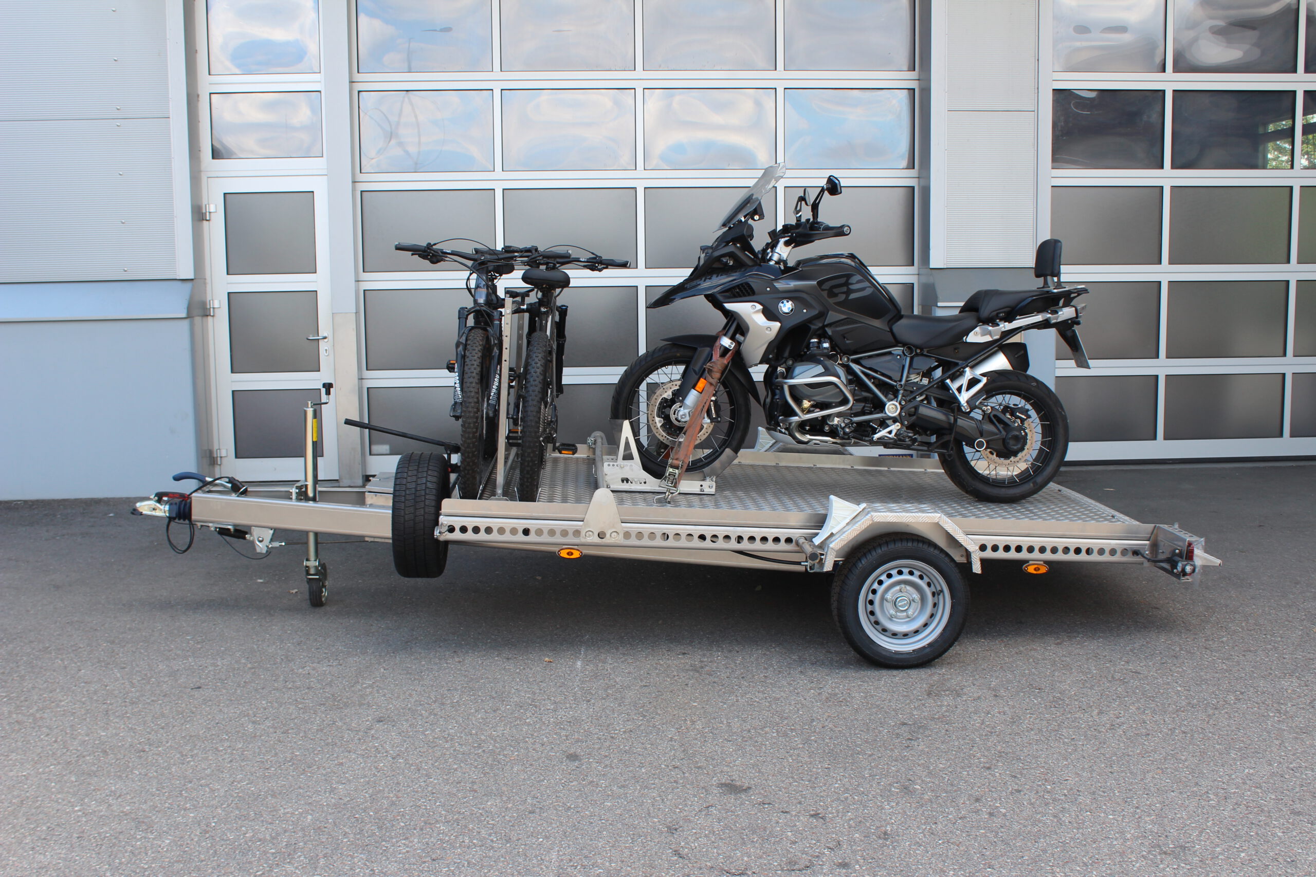 AS300 Aluminium Absenkanhänger in silber mit 2 E-Bikes auf der Plattform vorne quer beladen und mit einer BMW GS beladen vor einem Hallentor