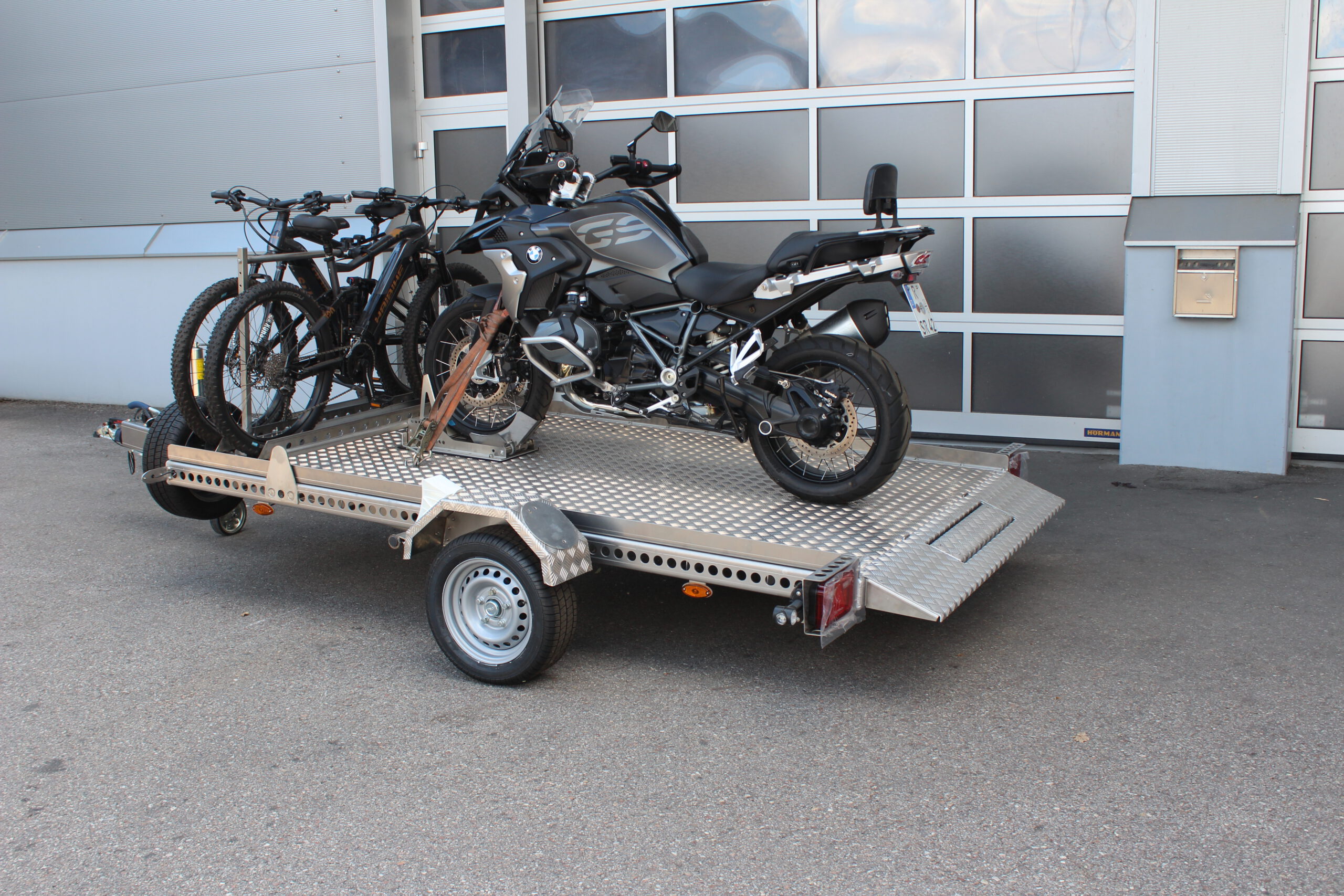 AS300 Aluminium Absenkanhänger in silber mit 2 E-Bikes auf der Plattform vorne quer beladen und mit einer BMW GS beladen vor einem Hallentor