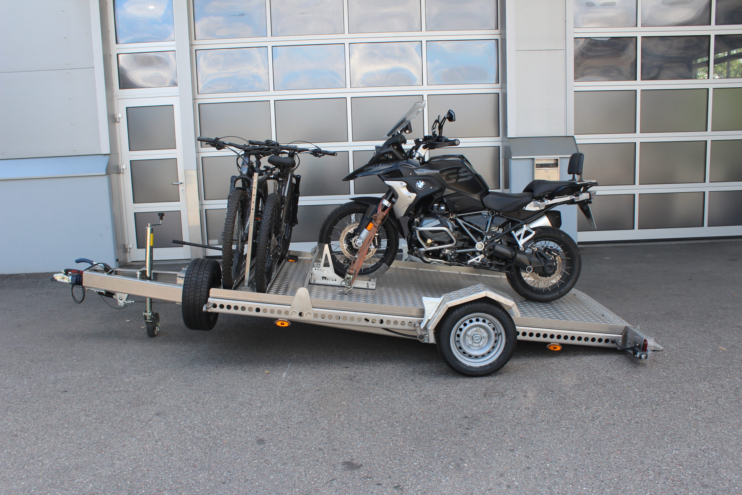 AS300 Aluminium Absenkanhänger in silber mit 2 E-Bikes auf der Plattform vorne quer beladen und mit einer BMW GS beladen vor einem Hallentor