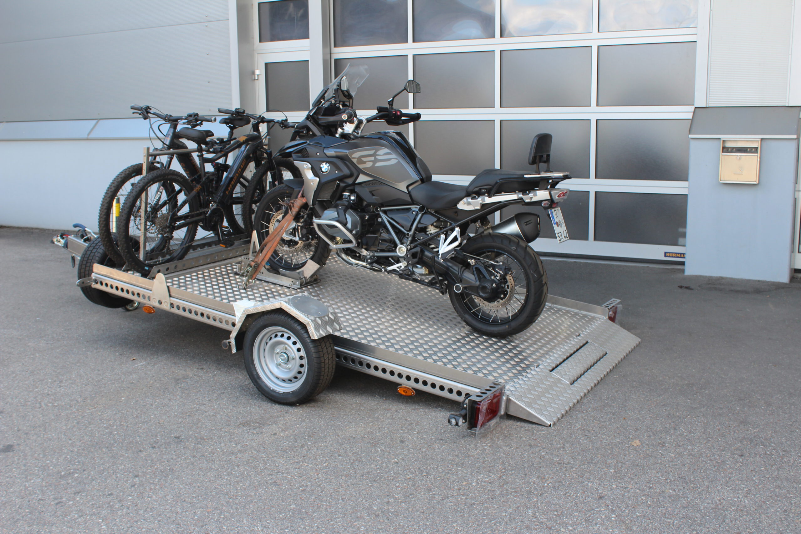AS300 Aluminium Absenkanhänger in silber mit 2 E-Bikes auf der Plattform vorne quer beladen und mit einer BMW GS beladen vor einem Hallentor