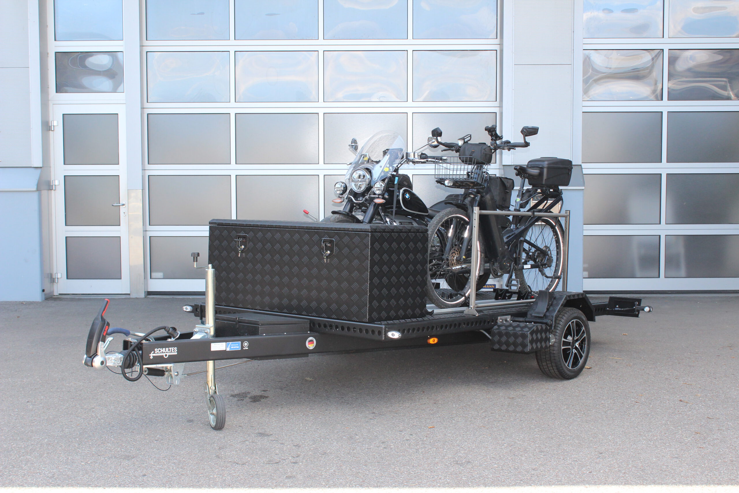Aluminium Absenkanhänger schwarz lackiert mit einer großen Staubox vorne auf der Plattform, mit einem Motorrad und einem Fahrrad beladen auf einem Hof vor einem Hallentor