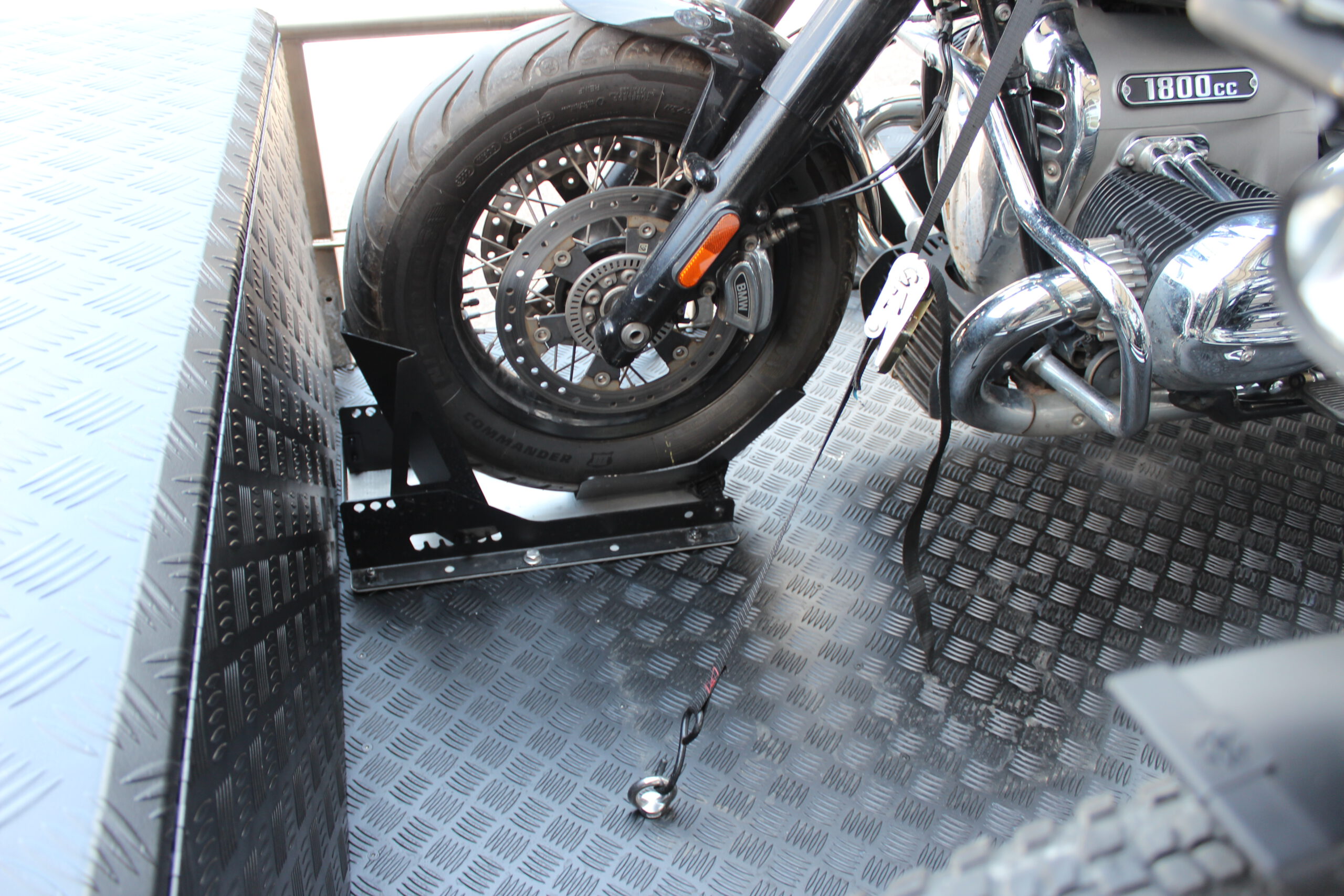 Aluminium Absenkanhänger schwarz lackiert mit einer großen Staubox vorne auf der Plattform, mit einem Motorrad und einem Fahrrad beladen auf einem Hof vor einem Hallentor