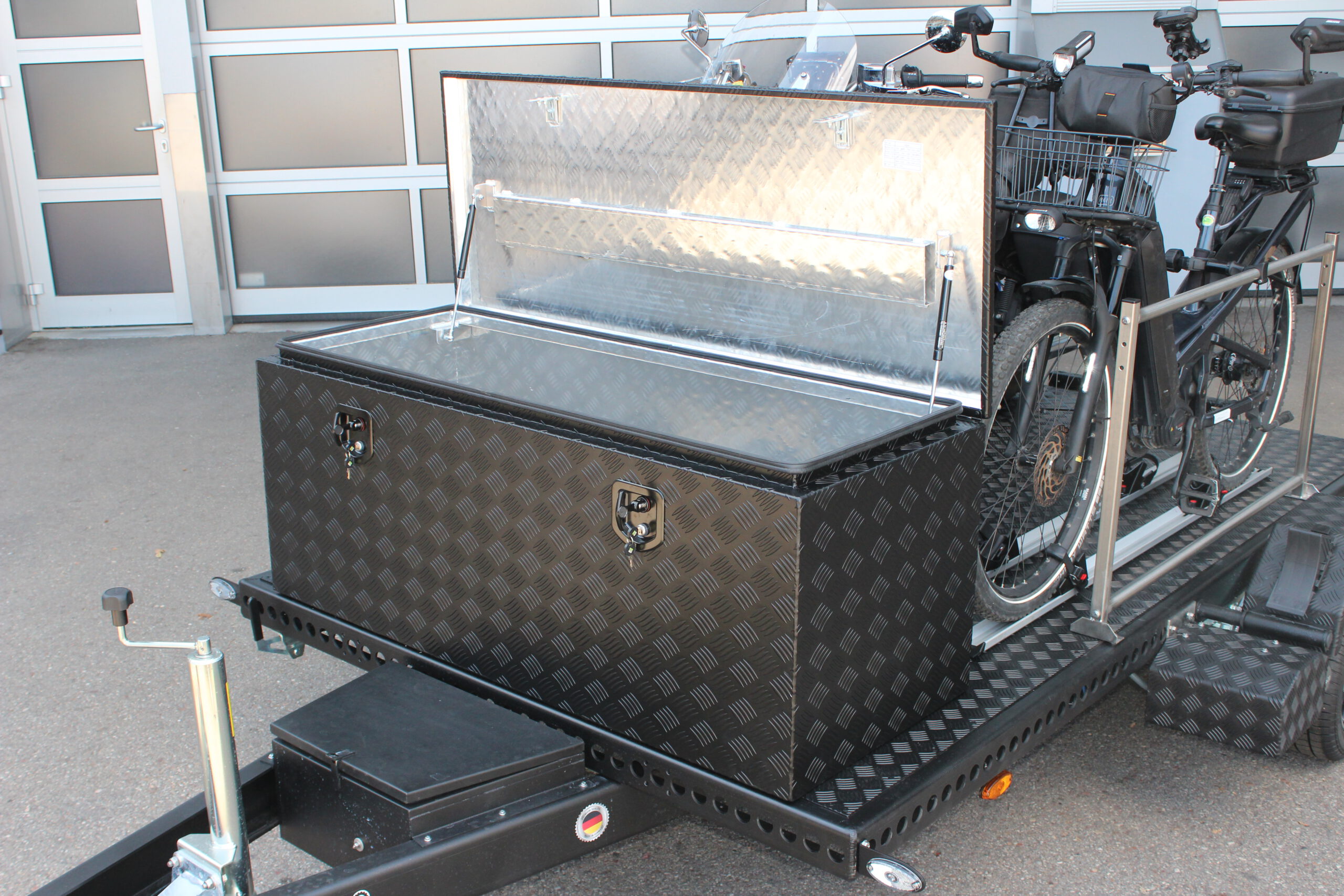 Aluminium Absenkanhänger schwarz lackiert mit einer großen Staubox vorne auf der Plattform, mit einem Motorrad und einem Fahrrad beladen auf einem Hof vor einem Hallentor