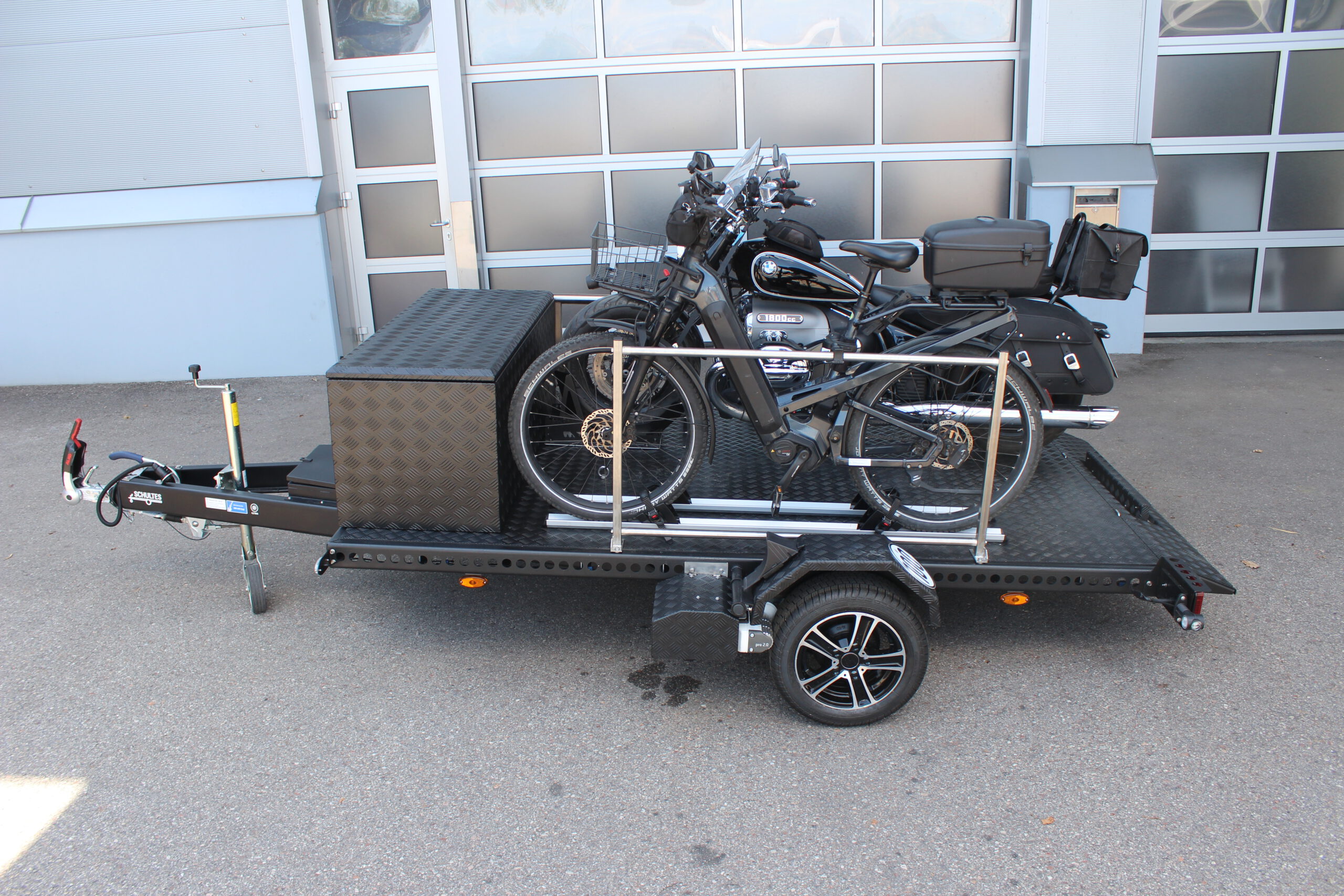 Aluminium Absenkanhänger schwarz lackiert mit einer großen Staubox vorne auf der Plattform, mit einem Motorrad und einem Fahrrad beladen auf einem Hof vor einem Hallentor