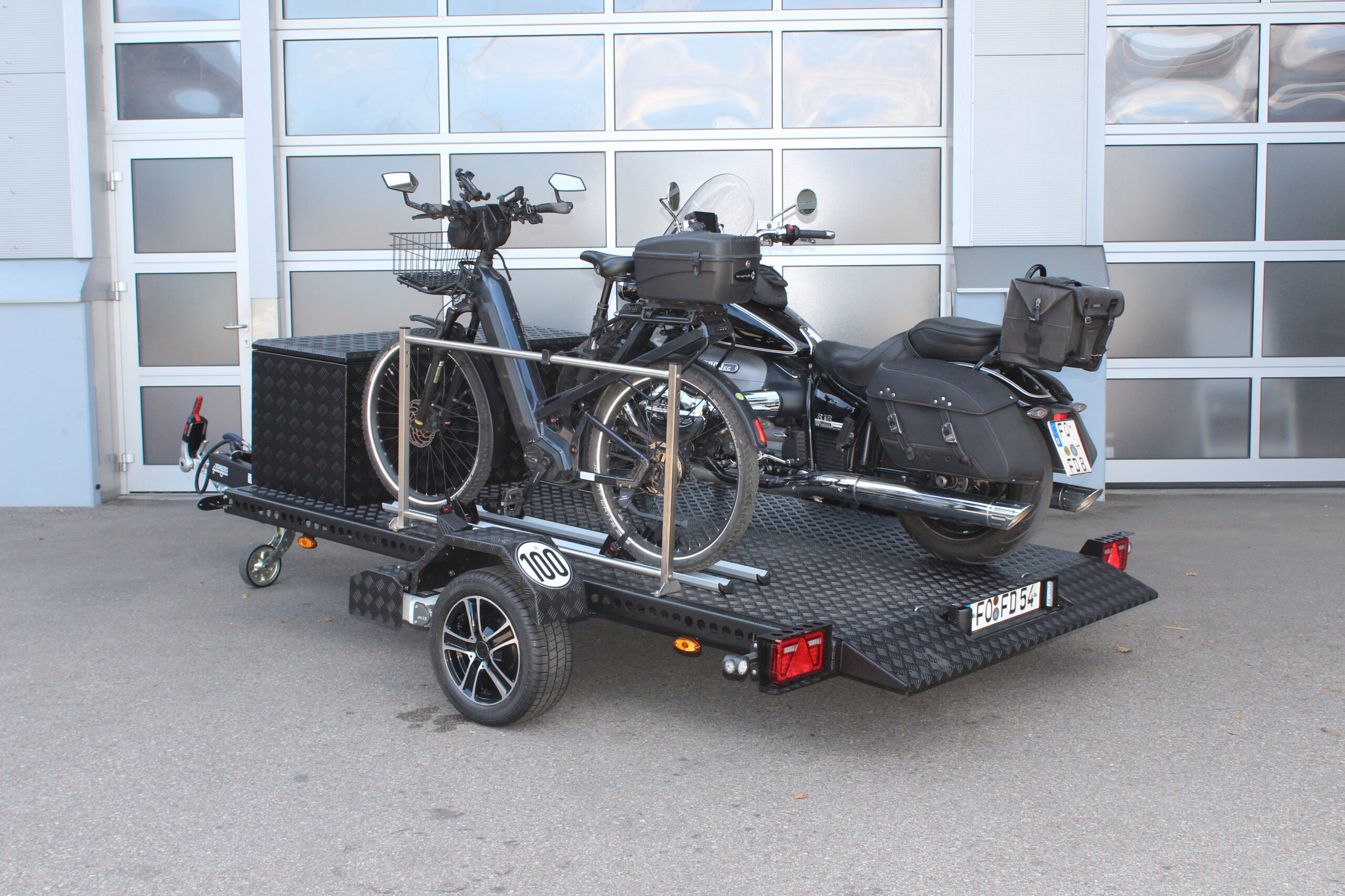 Aluminium Absenkanhänger schwarz lackiert mit einer großen Staubox vorne auf der Plattform, mit einem Motorrad und einem Fahrrad beladen auf einem Hof vor einem Hallentor