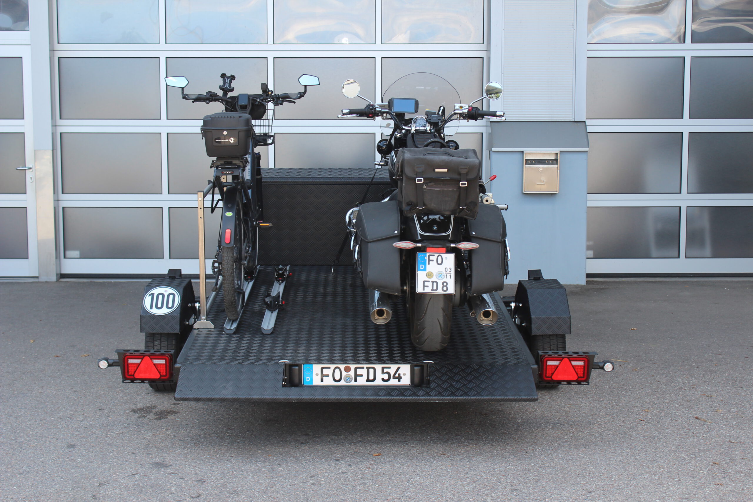 Aluminium Absenkanhänger schwarz lackiert mit einer großen Staubox vorne auf der Plattform, mit einem Motorrad und einem Fahrrad beladen auf einem Hof vor einem Hallentor
