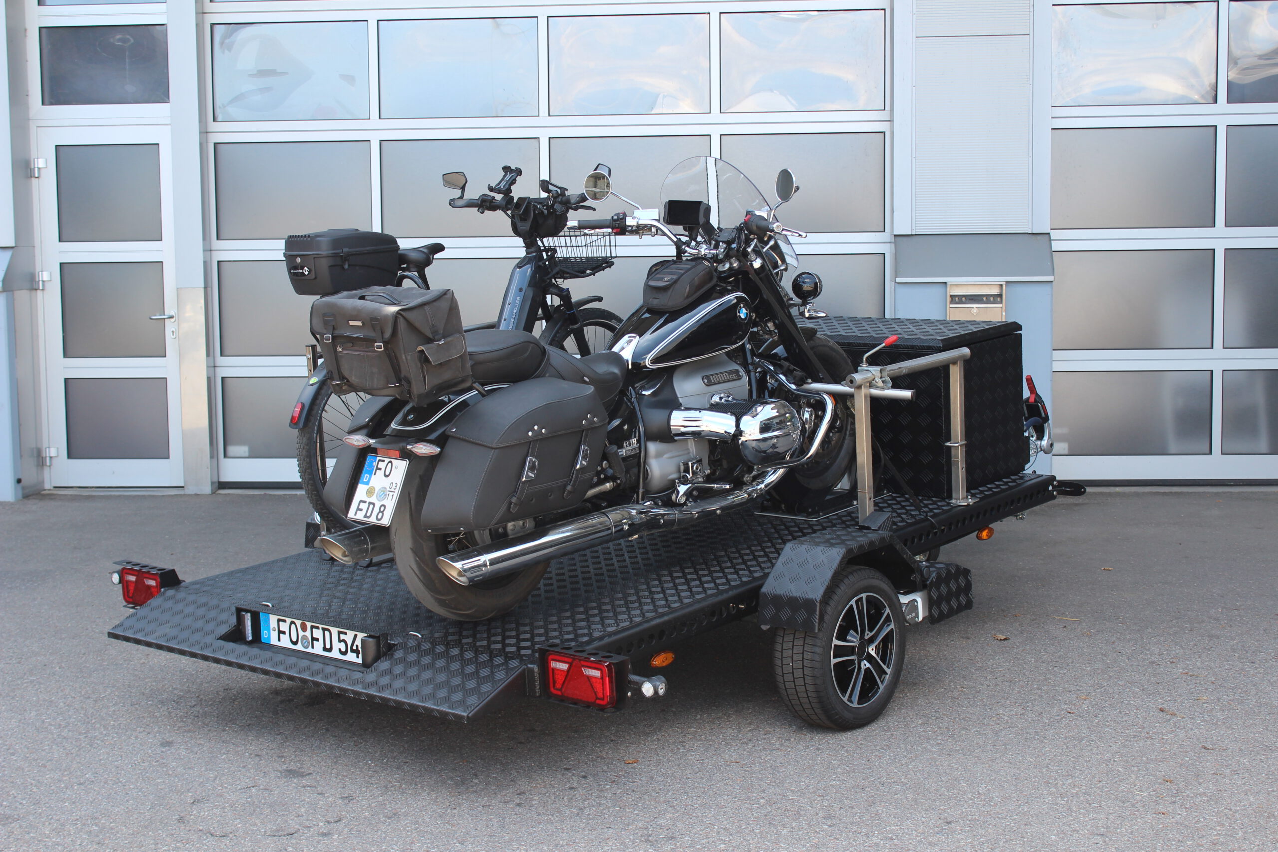 Aluminium Absenkanhänger schwarz lackiert mit einer großen Staubox vorne auf der Plattform, mit einem Motorrad und einem Fahrrad beladen auf einem Hof vor einem Hallentor