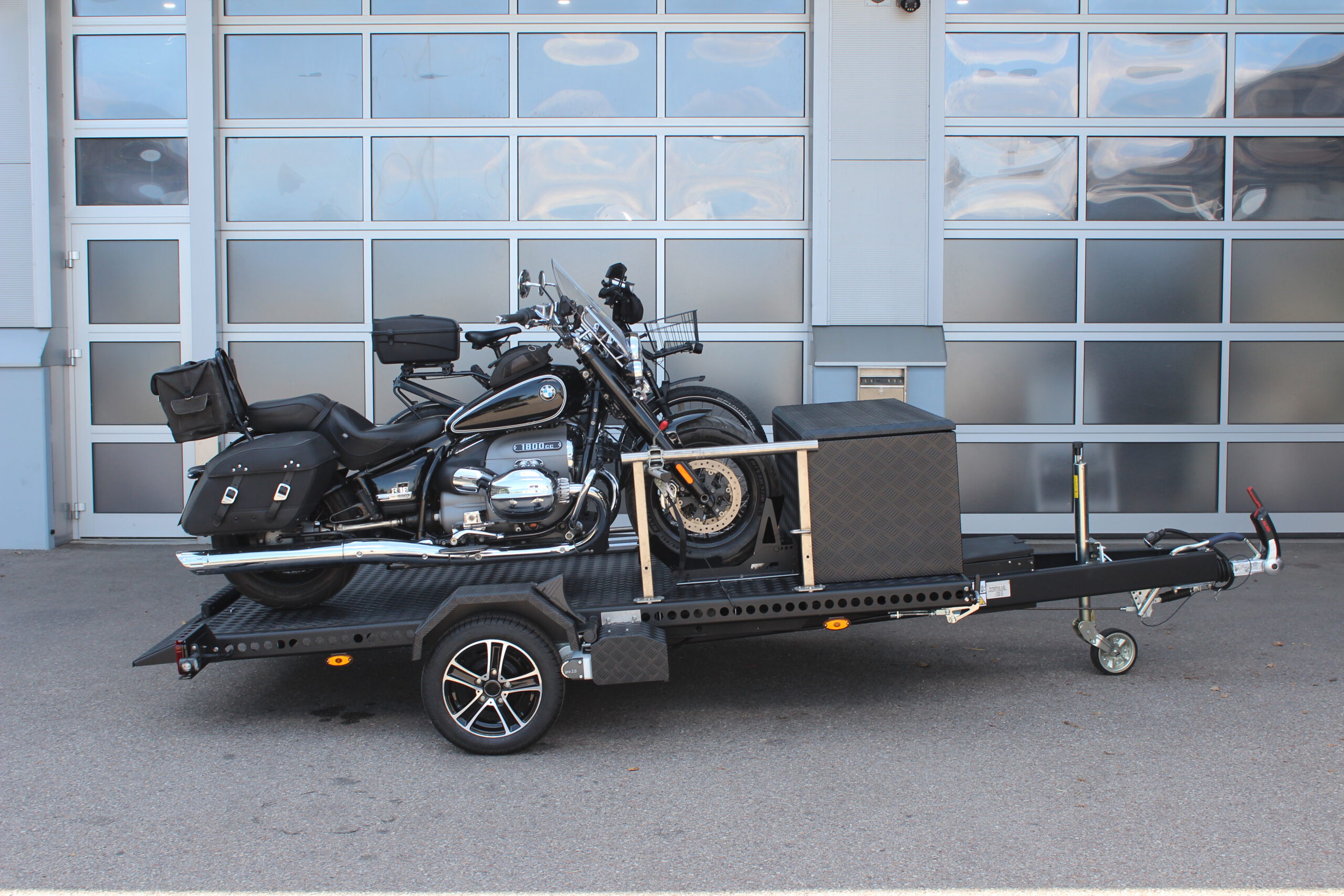Aluminium Absenkanhänger schwarz lackiert mit einer großen Staubox vorne auf der Plattform, mit einem Motorrad und einem Fahrrad beladen auf einem Hof vor einem Hallentor
