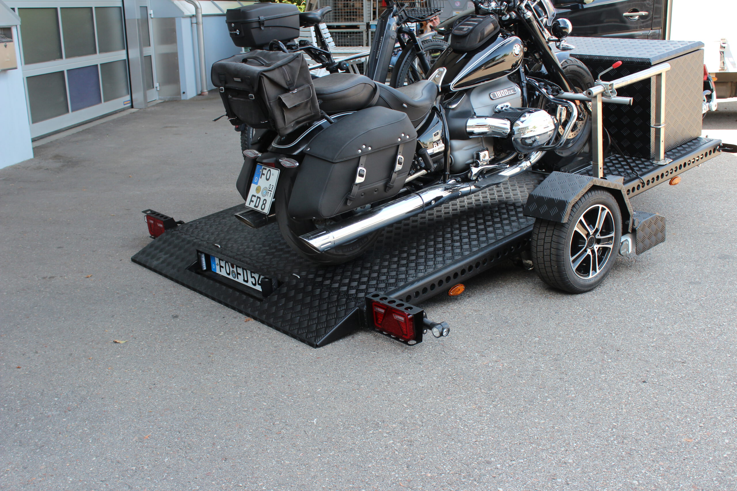 Aluminium Absenkanhänger schwarz lackiert mit einer großen Staubox vorne auf der Plattform, mit einem Motorrad und einem Fahrrad beladen auf einem Hof vor einem Hallentor