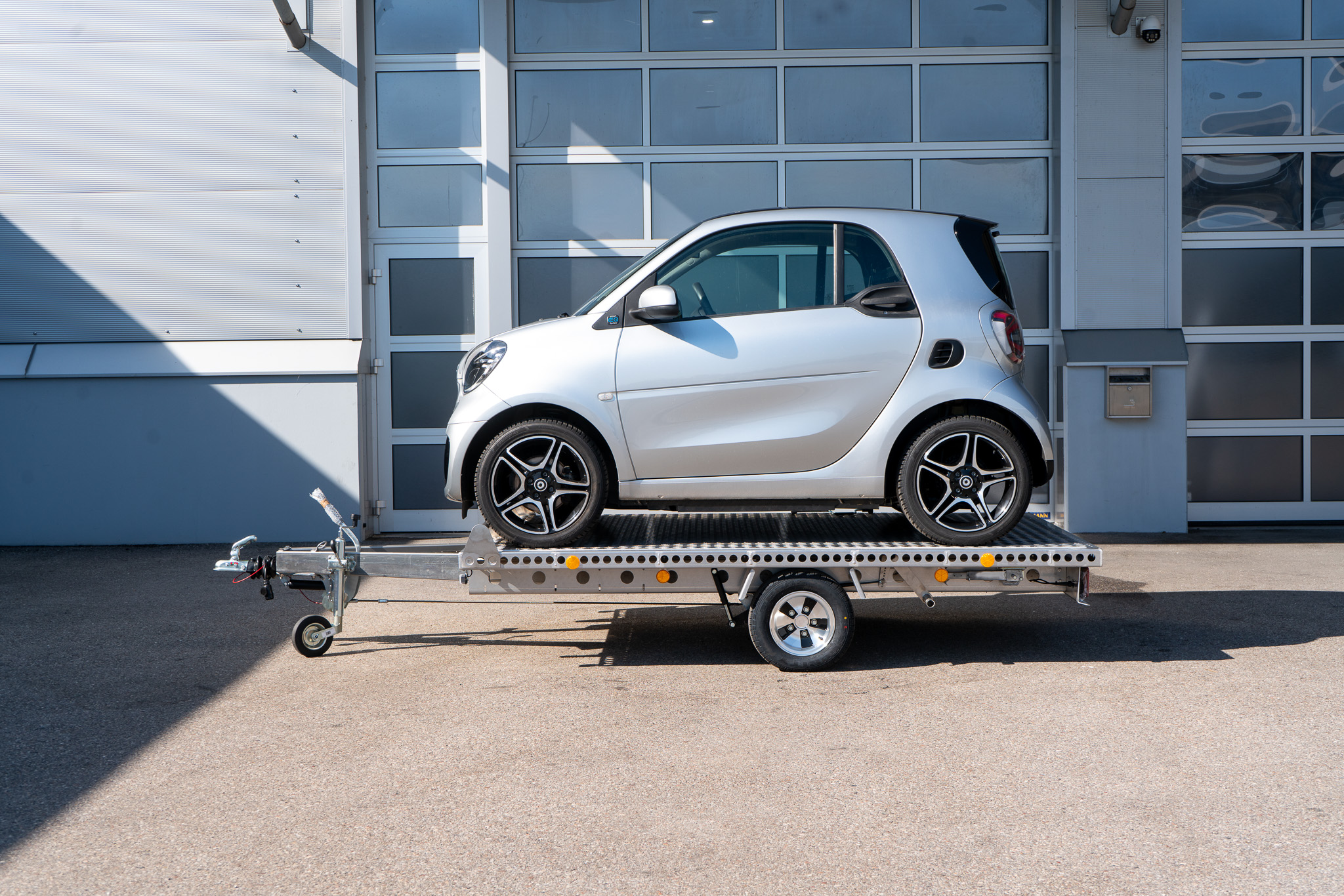 Alu Plattformanhänger mit zwei Auffahrrampen - Smart fortwo auf dem Anhänger, vor einem Hallentor - Seitenansicht