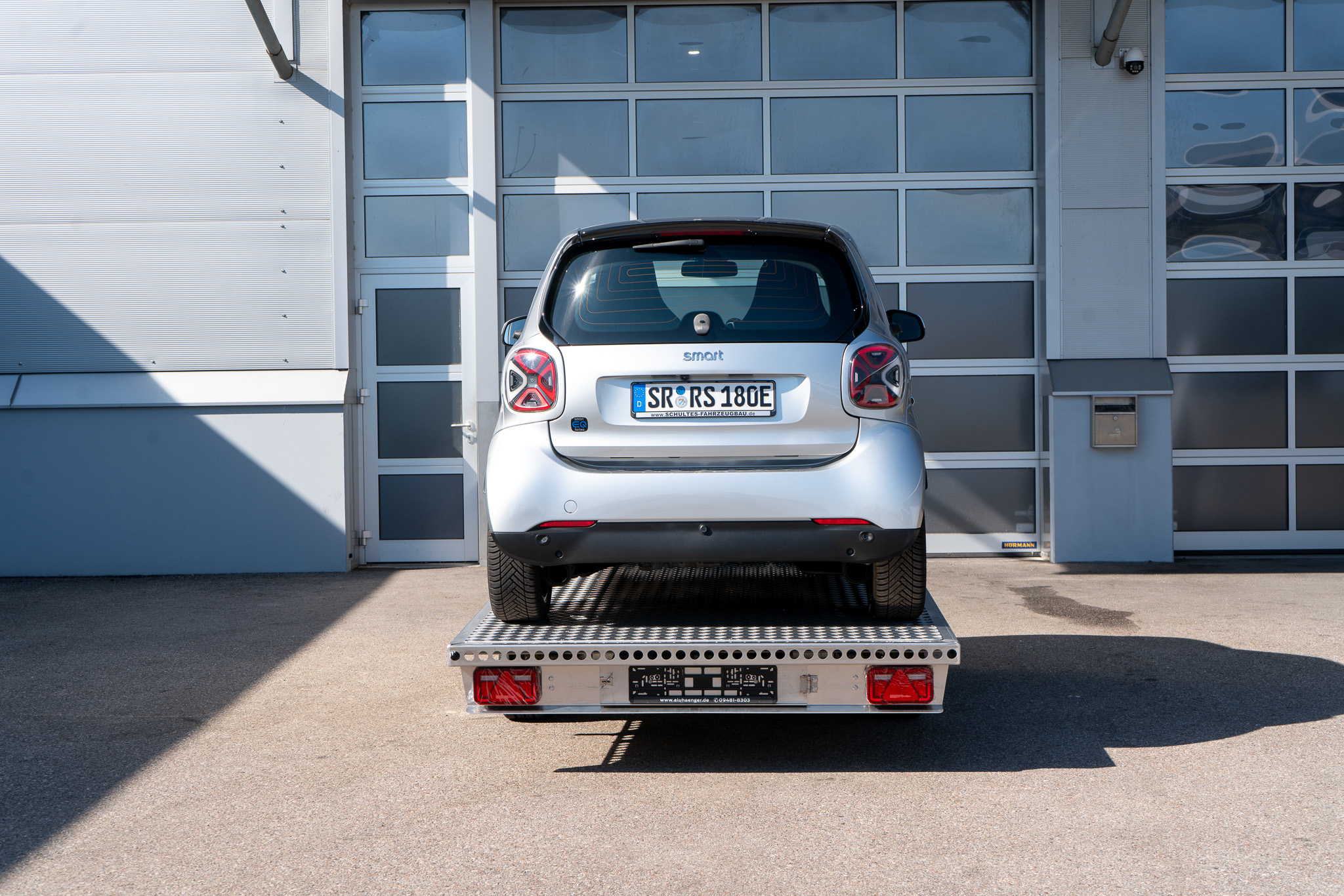 Alu Plattformanhänger mit zwei Auffahrrampen - Smart fortwo auf dem Anhänger, vor einem Hallentor - Heckansicht