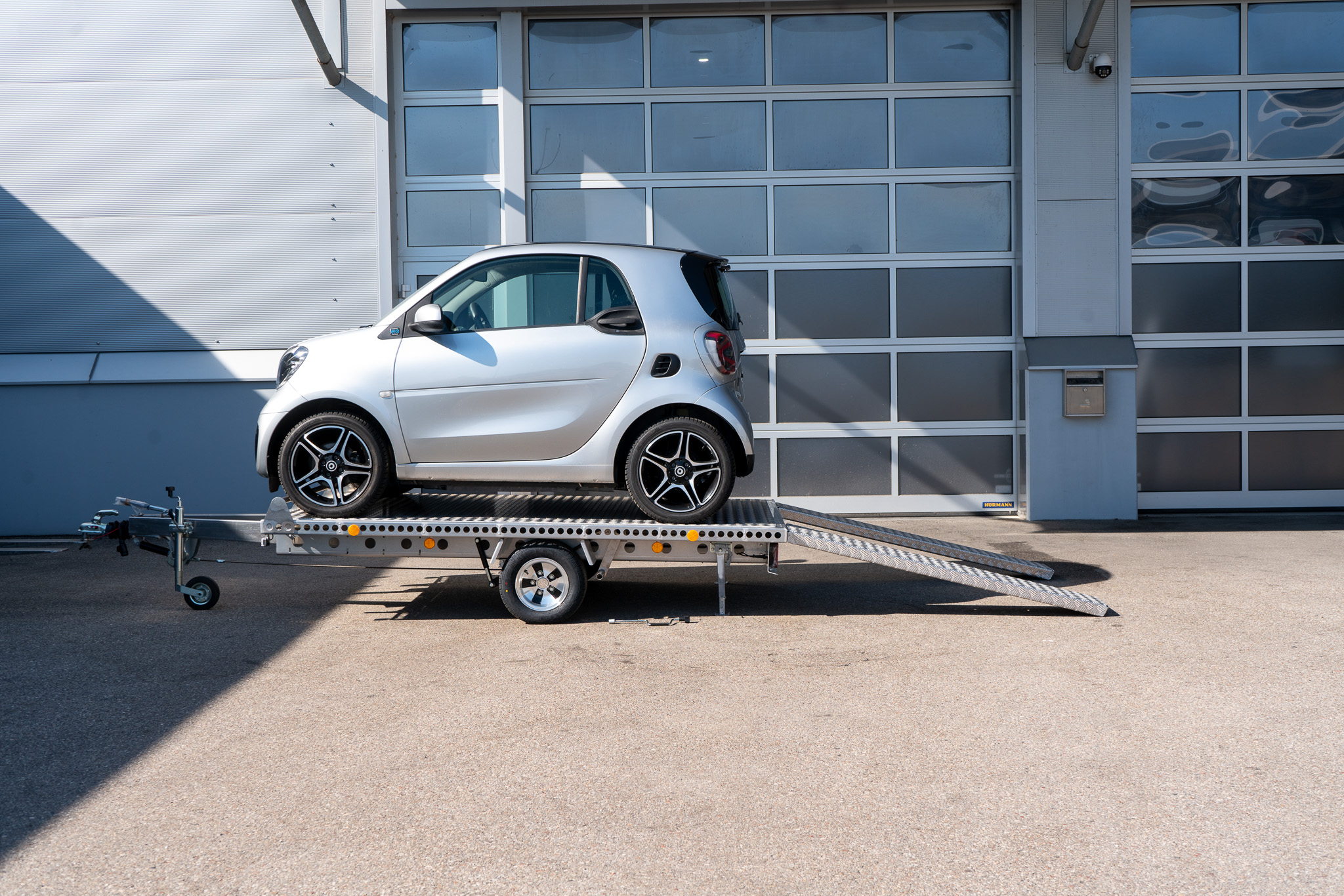 Alu Plattformanhänger mit zwei Auffahrrampen - Smart fortwo auf dem Anhänger, vor einem Hallentor - Seitenansicht