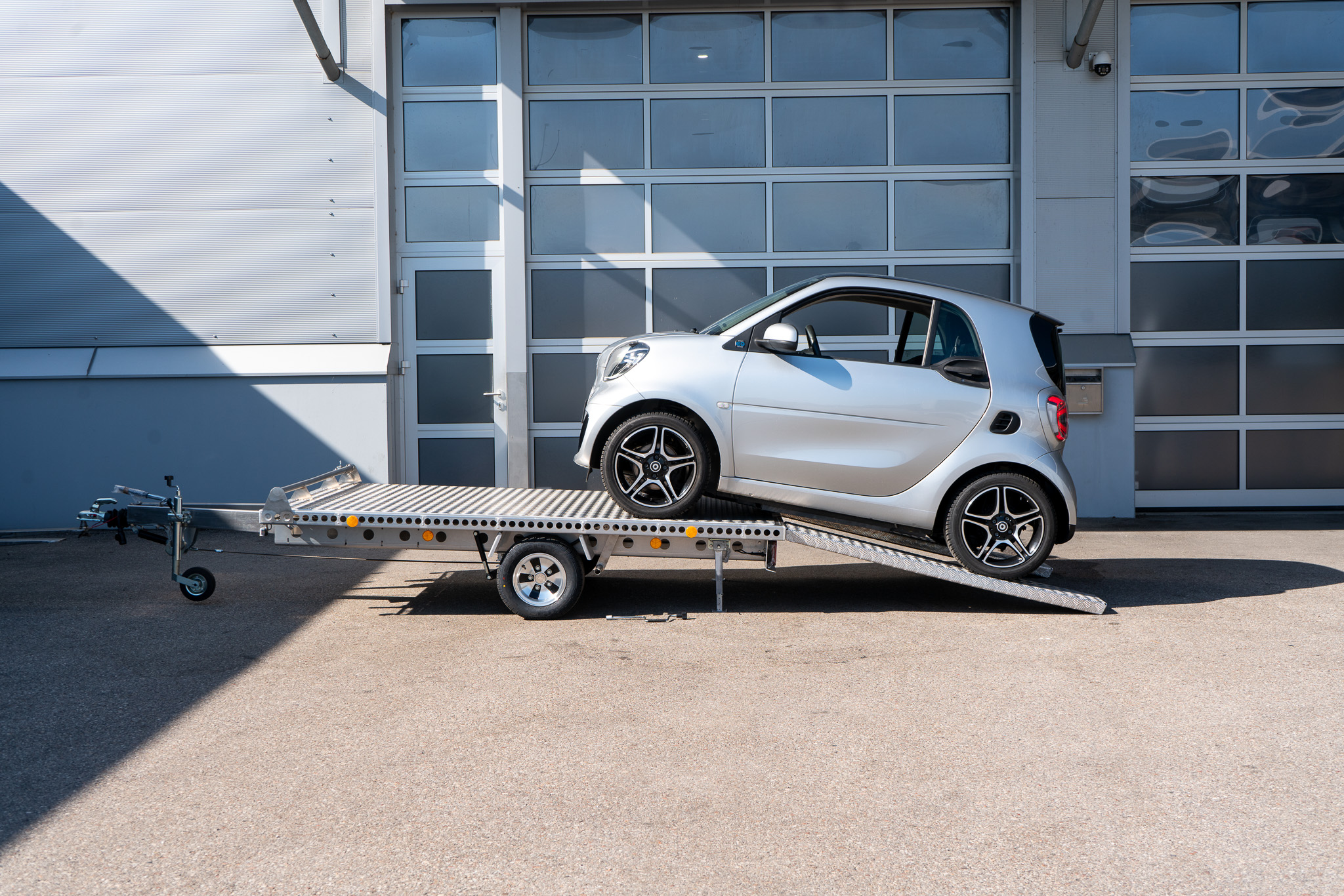 Alu Plattformanhänger mit zwei Auffahrrampen auf denen ein Smart fortwo auf den Anhänger fährt, vor einem Hallentor - Seitenansicht