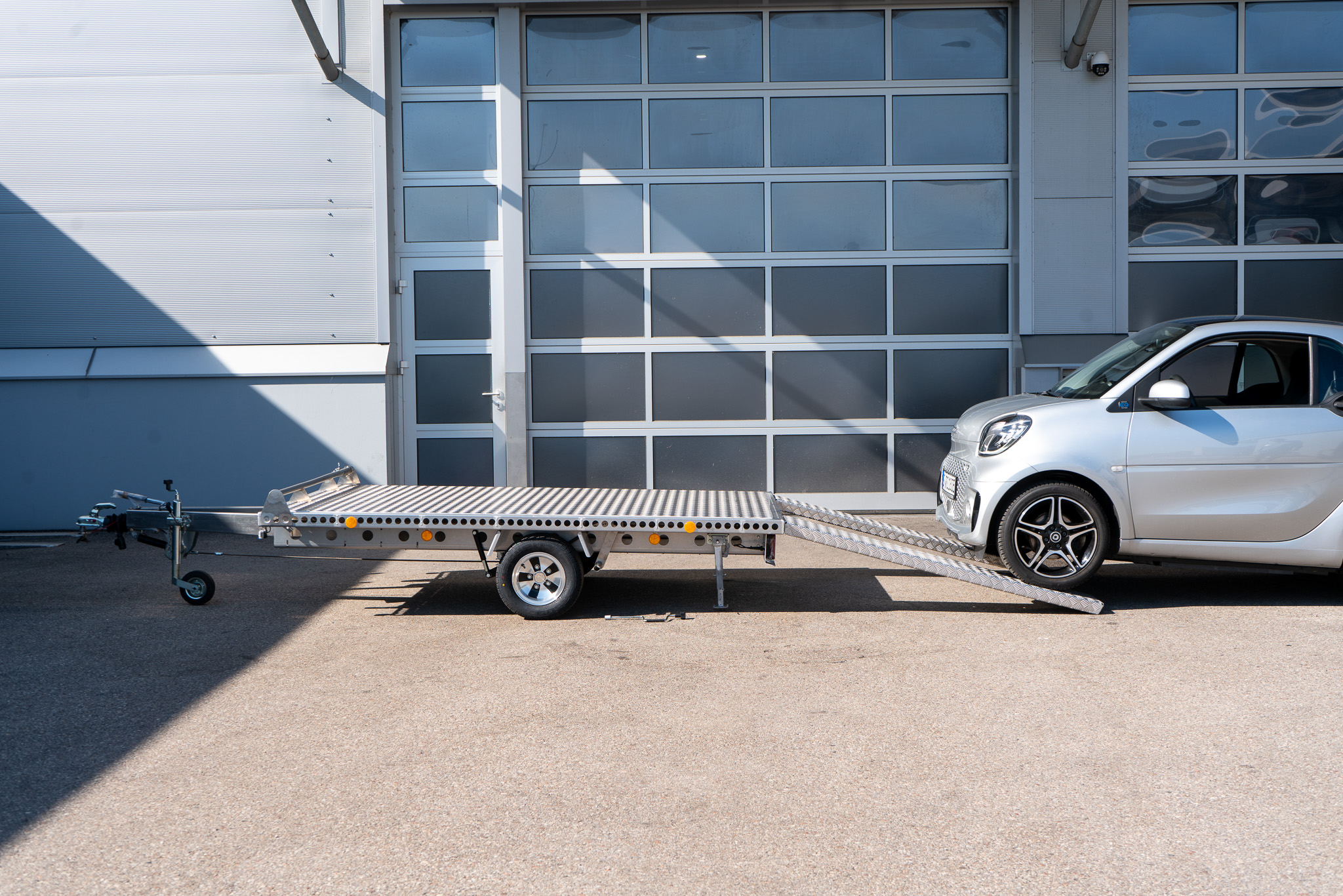 Alu Plattformanhänger mit zwei Auffahrrampen auf denen ein Smart fortwo auf den Anhänger fährt, vor einem Hallentor - Seitenansicht